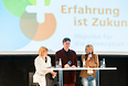 Sabine Heimbach, Dominik Schott und Uschi Glas an einem Tisch auf der Hauptbühne der Messe "Die 66", im Hintergrund eingeblendet das Logo von "Erfahrung ist Zukunft"